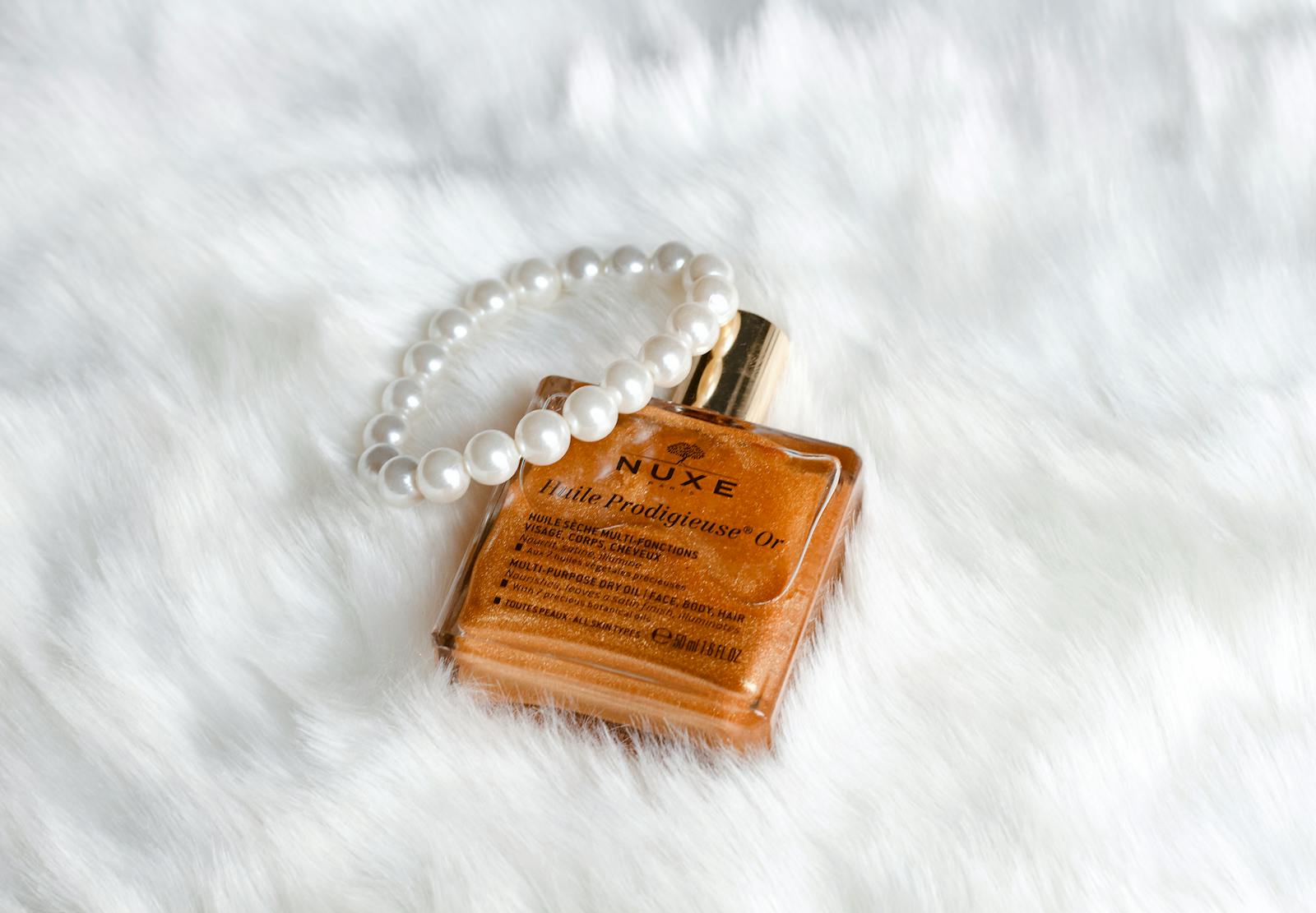 From above golden face oil in refined bottle placed with pearl bracelet on white cozy plaid