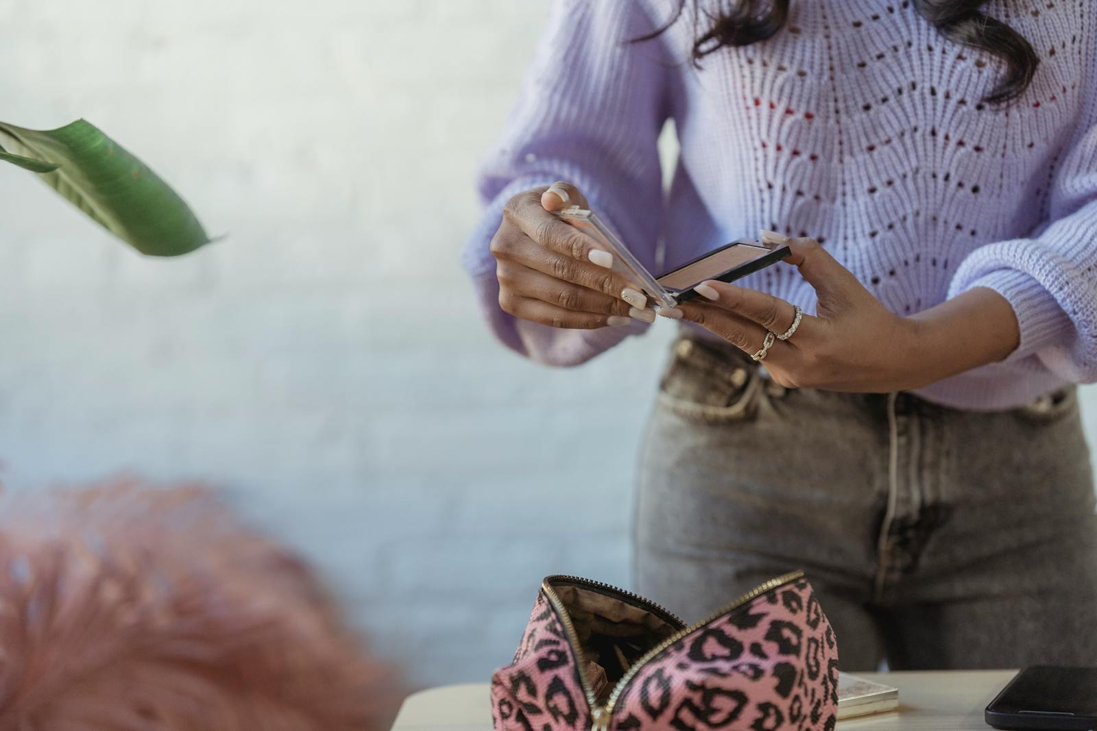 Crop anonymous African American female opening mirror at table with cosmetic bag on blurred background